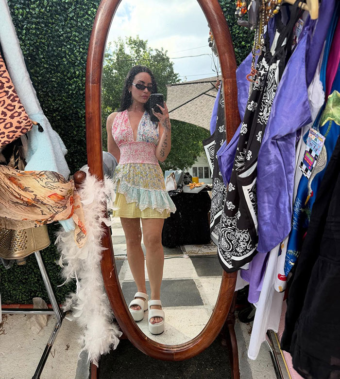 Person in summer dress and sunglasses taking a mirror selfie, surrounded by colorful clothing. Person in summer dress and sunglasses taking a mirror selfie, surrounded by colorful clothing.