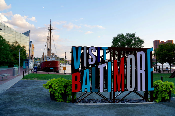 Baltimore cityscape with a prominent "Visit Baltimore" sign under a clear sky, near a historic ship. Baltimore cityscape with a prominent "Visit Baltimore" sign under a clear sky, near a historic ship.