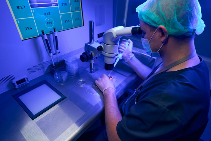 Scientist in lab using microscope for fertility research, wearing blue scrubs and hairnet. Scientist in lab using microscope for fertility research, wearing blue scrubs and hairnet.