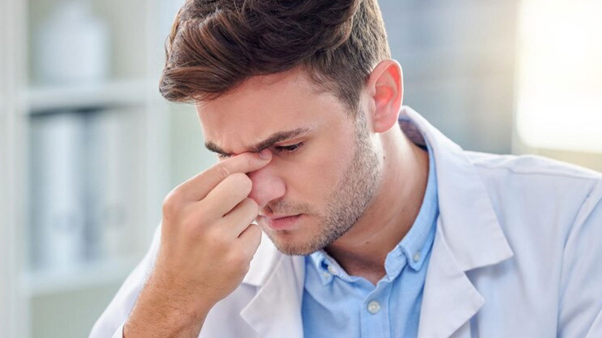 Young male doctor in white coat looking stressed while pinching bridge of nose indoors with blurred background medical questions concept