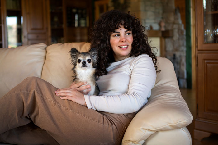 A woman with curly hair lovingly holds a small dog on a beige sofa, highlighting emotional support and hope. A woman with curly hair lovingly holds a small dog on a beige sofa, highlighting emotional support and hope.
