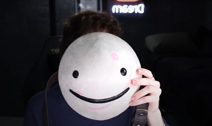 Youtuber Dream holding a large smiley face mask covering their face with Dream sign glowing in the background, related to Dream face reveal.