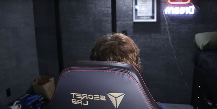 Youtuber Dream sitting in a Secret Lab gaming chair facing away, with a Dream neon sign visible in a dimly lit room.