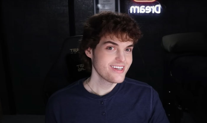 Youtuber Dream with curly hair smiling in a dark room with a glowing Dream sign, representing Minecraft star’s face reveal moment.