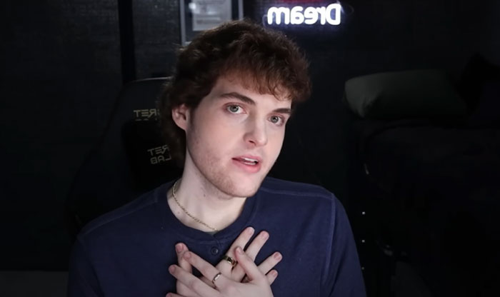 Youtuber Dream with curly hair sitting in a dark room with a glowing Dream sign, discussing the Dream face reveal and fan reactions.