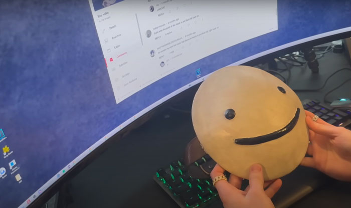 Youtuber Dream holding a round mask with a simple smiley face in front of a curved computer screen displaying comments.