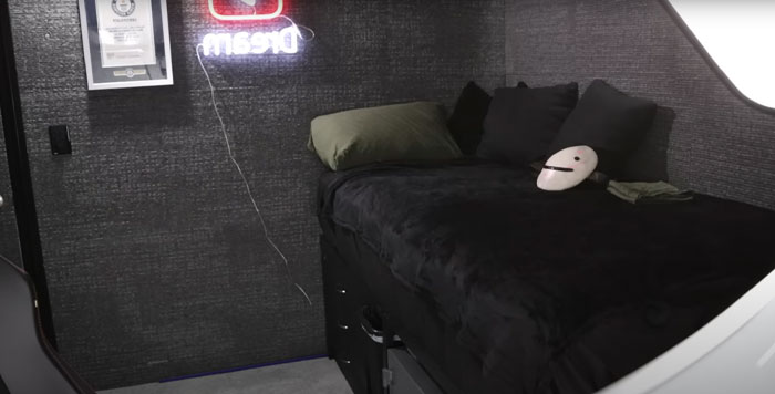Small bedroom with black bedding and pillows, featuring a Dream face mask and neon sign related to Minecraft star.