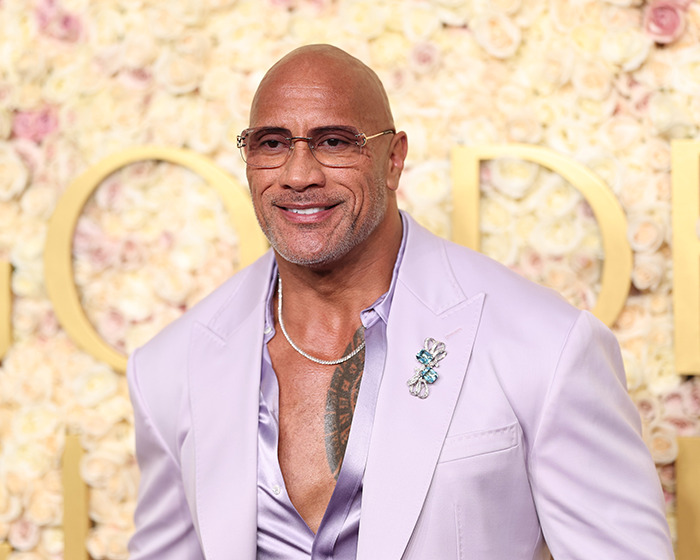 Bald man in a lavender suit with glasses and a floral backdrop, sparking race confusion online. Bald man in a lavender suit with glasses and a floral backdrop, sparking race confusion online.