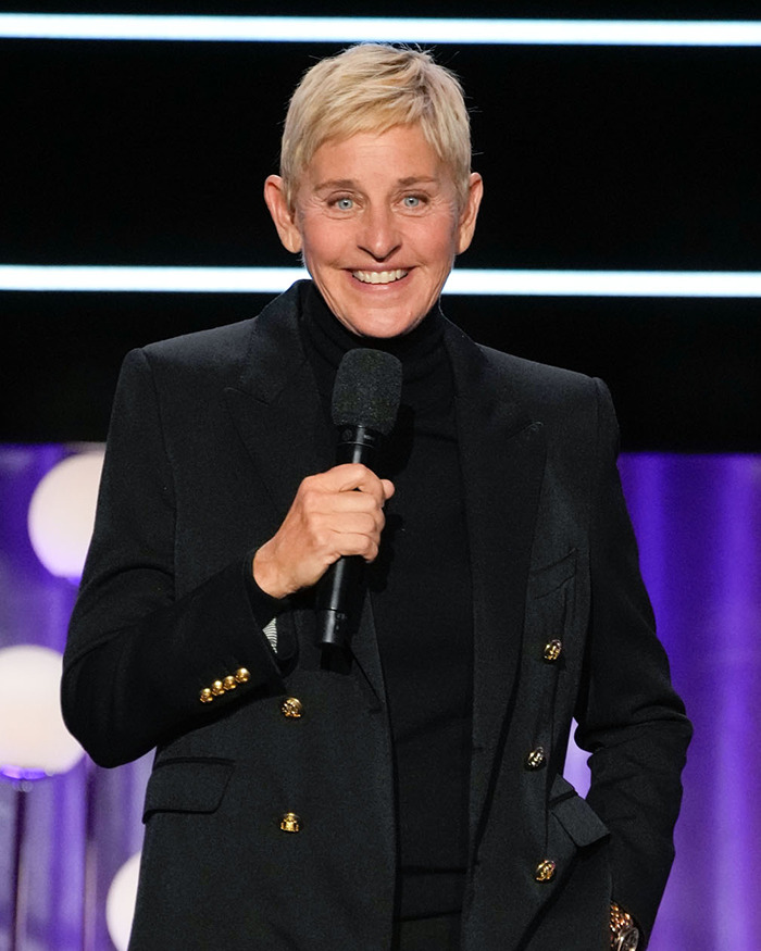 Ellen DeGeneres holding a microphone, wearing a black suit, smiling on stage. Ellen DeGeneres holding a microphone, wearing a black suit, smiling on stage.