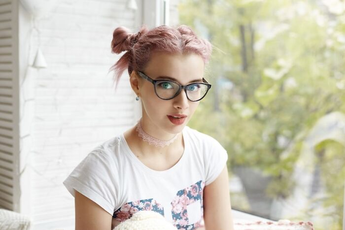 Young woman with pink hair and glasses sits by a window, embodying a reflective mood.