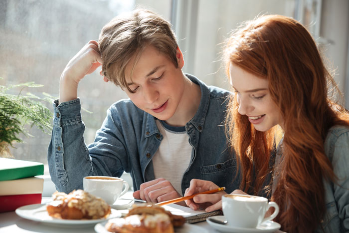 Teen couple smiling at a table with coffee, disregarding dad's ban due to grades slipping. Teen couple smiling at a table with coffee, disregarding dad's ban due to grades slipping.