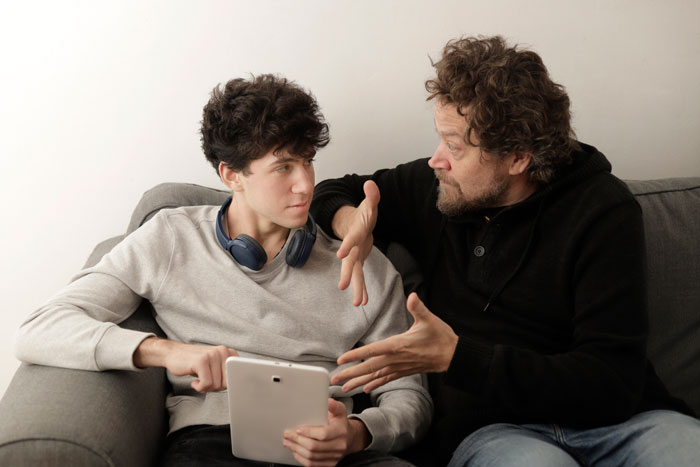 Father discusses grades with son holding a tablet, illustrating tension over son's girlfriend. Father discusses grades with son holding a tablet, illustrating tension over son's girlfriend.