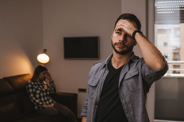 Man in a distressed pose in living room, with woman on sofa, discussing son's grades and relationship issues. Man in a distressed pose in living room, with woman on sofa, discussing son's grades and relationship issues.