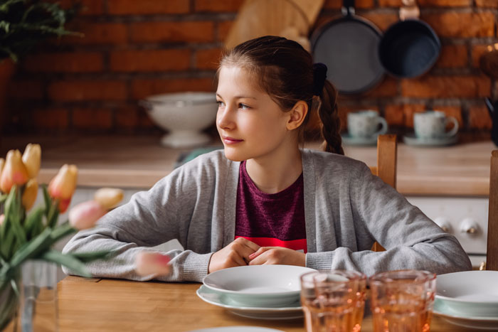 A young girl, possibly the daughter, sitting at a table set with plates and glasses, looking towards the side. A young girl, possibly the daughter, sitting at a table set with plates and glasses, looking towards the side.