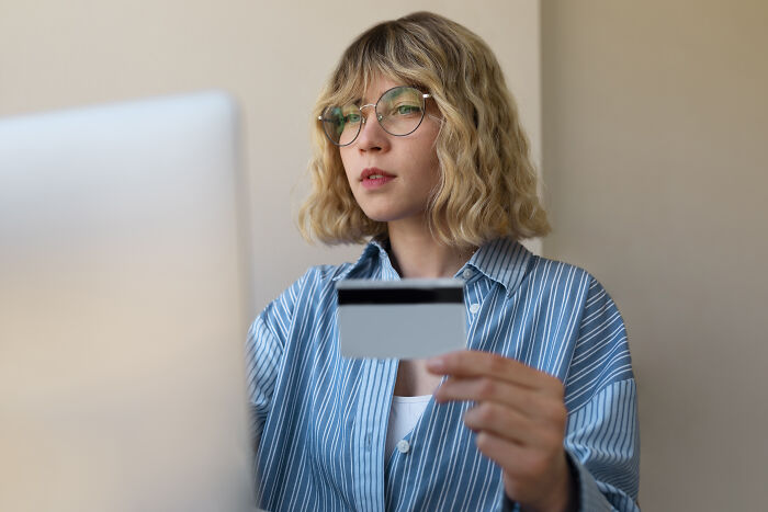 Woman holding credit card, wearing striped shirt and glasses, using a computer, possibly shopping online or managing finances.
