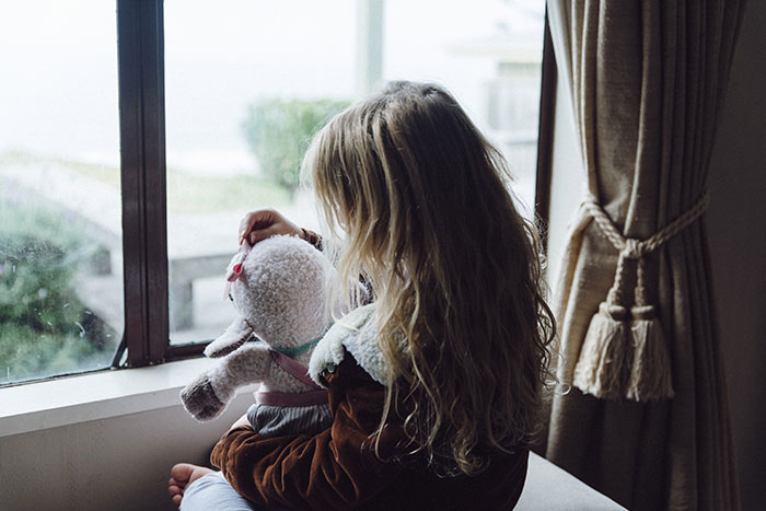 Young girl holding a stuffed toy, gazing out of a window, symbolizing waiting and uncertainty. Young girl holding a stuffed toy, gazing out of a window, symbolizing waiting and uncertainty.