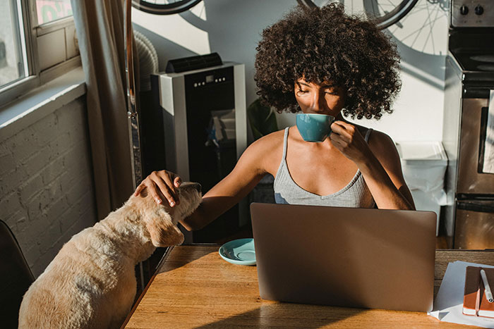 Woman drinking coffee at a table, petting a dog, with a laptop, reflecting casual birthday planning and diet challenges. Woman drinking coffee at a table, petting a dog, with a laptop, reflecting casual birthday planning and diet challenges.