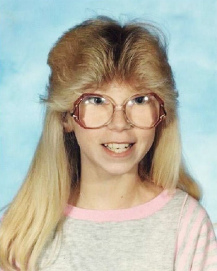 Blonde person with a retro hairstyle and large glasses, wearing a pink-striped shirt.