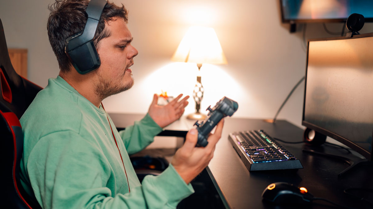Man wearing headphones holding a game controller, frustrated while sitting at a gaming setup with keyboard and monitor.
