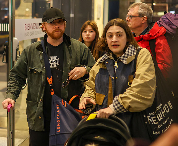 A man and a woman with luggage at a station, the man wearing a cap and jacket, the woman pushing a stroller. A man and a woman with luggage at a station, the man wearing a cap and jacket, the woman pushing a stroller.