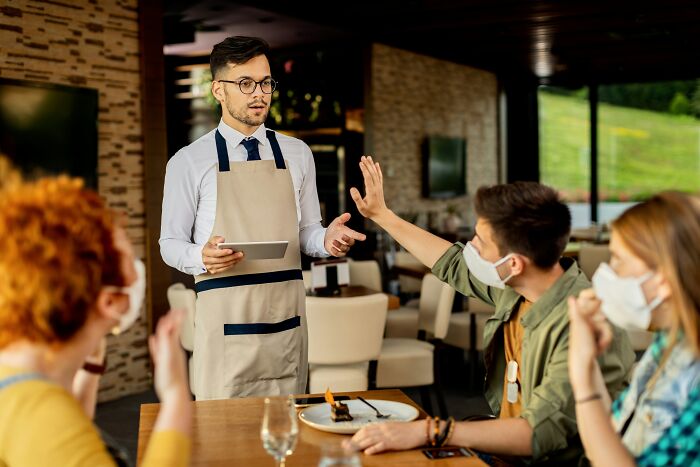 A waiter with a tablet engaging with masked diners at a restaurant, illustrating social interaction skills.