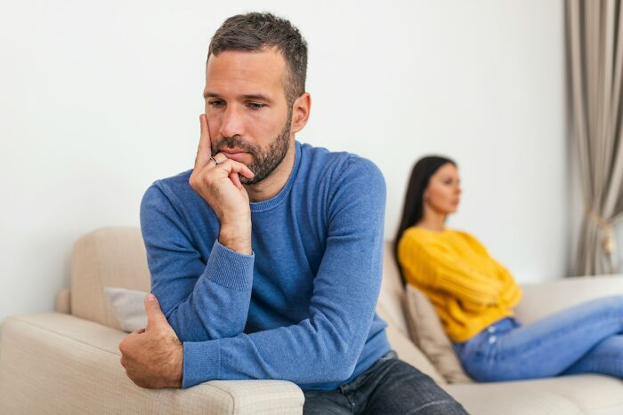 Man in blue sweater looking thoughtful, sitting on a couch with a woman in yellow sweater in the background.