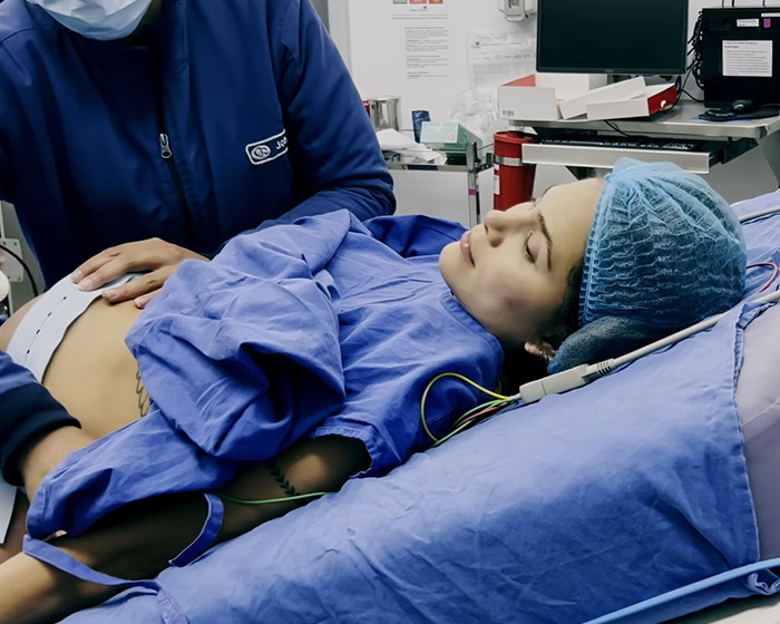 A woman in a hospital bed post-birth, attended by a doctor, wearing blue medical attire and cap. A woman in a hospital bed post-birth, attended by a doctor, wearing blue medical attire and cap.