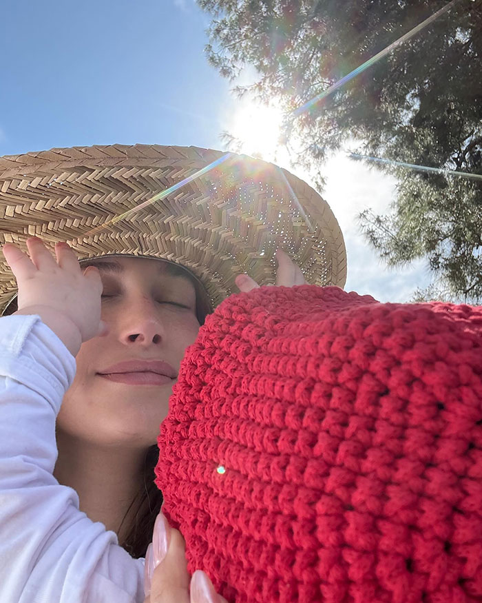 Woman in sun hat holding baby with red crochet blanket by the pool, sunlight streaming through trees. Keywords: Internet debate. Woman in sun hat holding baby with red crochet blanket by the pool, sunlight streaming through trees. Keywords: Internet debate.