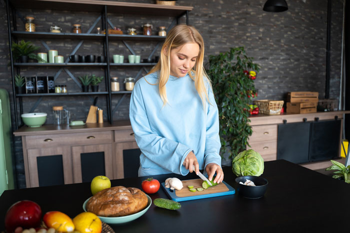 Woman in blue sweatshirt slicing cucumber, standing in a modern kitchen with various vegetables on the counter. Woman in blue sweatshirt slicing cucumber, standing in a modern kitchen with various vegetables on the counter.