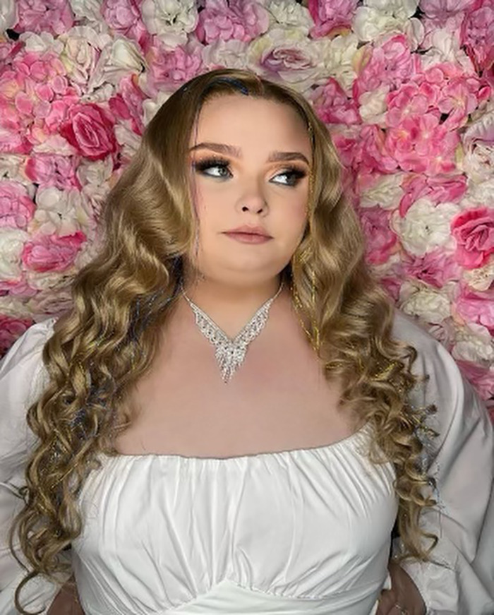 Young woman with long, curly hair in front of a floral backdrop, wearing a white dress and necklace. Young woman with long, curly hair in front of a floral backdrop, wearing a white dress and necklace.