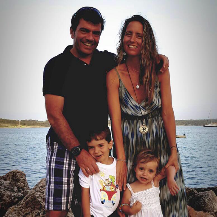 A smiling family of four stands outdoors near the water, with a sailboat visible in the background. A smiling family of four stands outdoors near the water, with a sailboat visible in the background.