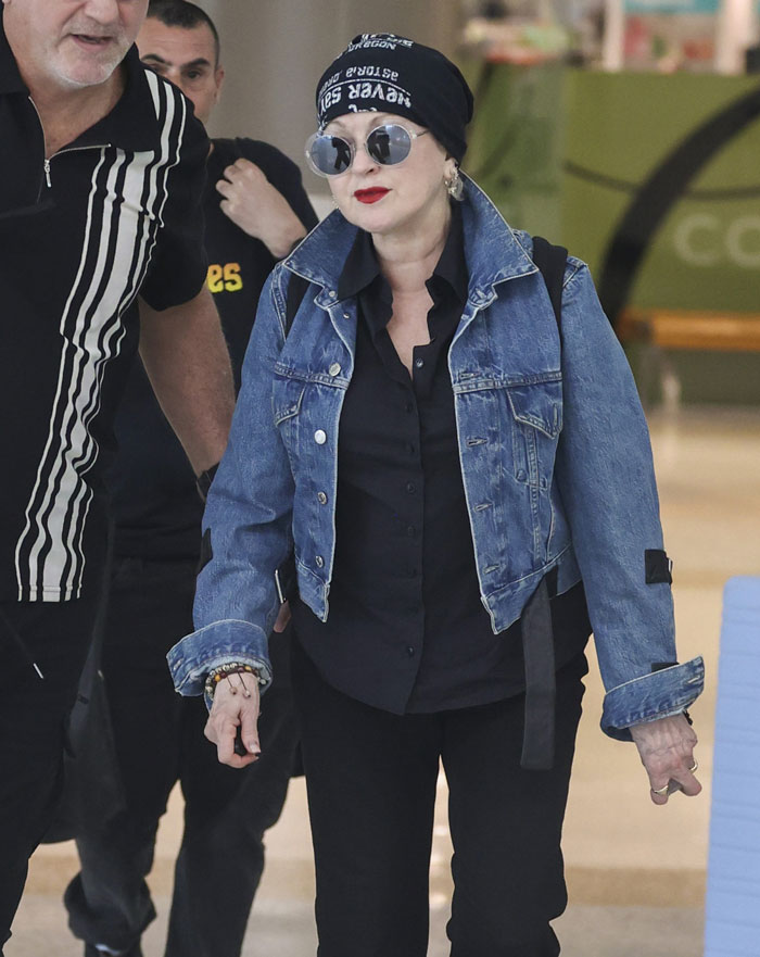 Cyndi Lauper appears unrecognizable, wearing a denim jacket, scarf, and sunglasses in a casual setting.