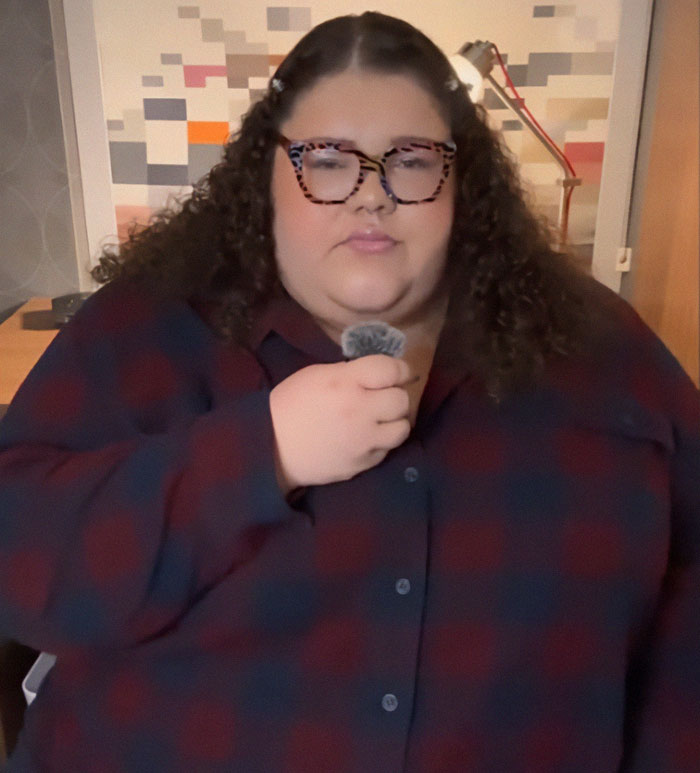 Plus-sized passenger with glasses holding a microphone, expressing concern in an indoor setting. Plus-sized passenger with glasses holding a microphone, expressing concern in an indoor setting.