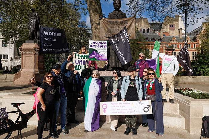 Protesters with signs on trans women and legal status outside a court, standing near a statue. Protesters with signs on trans women and legal status outside a court, standing near a statue.