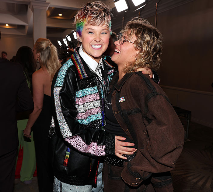 JoJo Siwa smiling at an event, wearing a colorful jacket, with a friend in a lively atmosphere. JoJo Siwa smiling at an event, wearing a colorful jacket, with a friend in a lively atmosphere.
