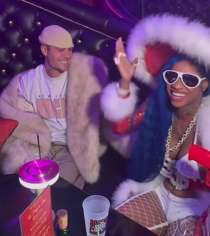 Celebrity smiling beside a female rapper in vibrant attire at a lively club setting. Celebrity smiling beside a female rapper in vibrant attire at a lively club setting.