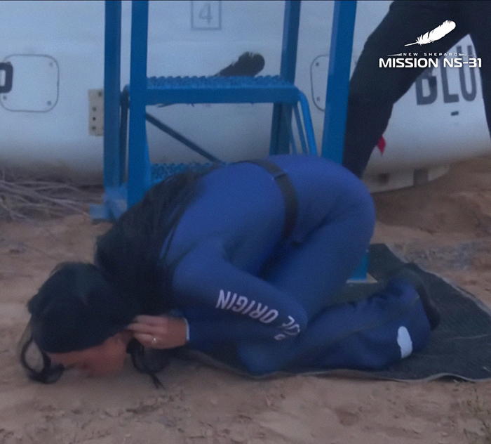 Person in blue space suit kneeling on the ground, with "New Shepard" and "Mission NS-31" visible, showing respect. Person in blue space suit kneeling on the ground, with "New Shepard" and "Mission NS-31" visible, showing respect.