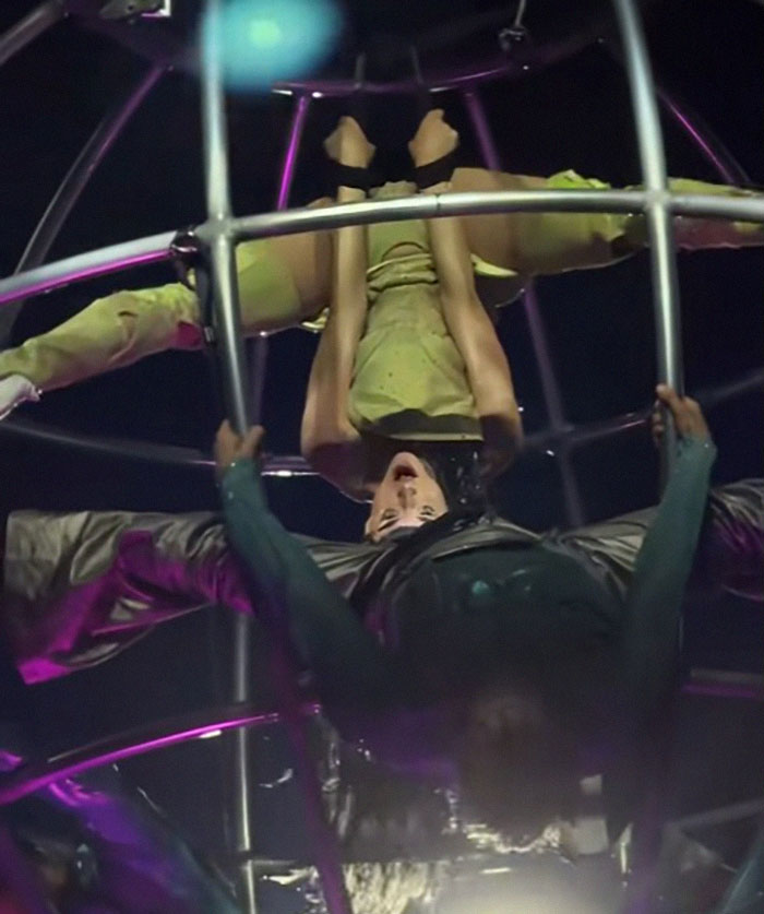 Singer performs a daring move upside down in a metal contraption during a tour, showcasing energetic dance choreography. Singer performs a daring move upside down in a metal contraption during a tour, showcasing energetic dance choreography.