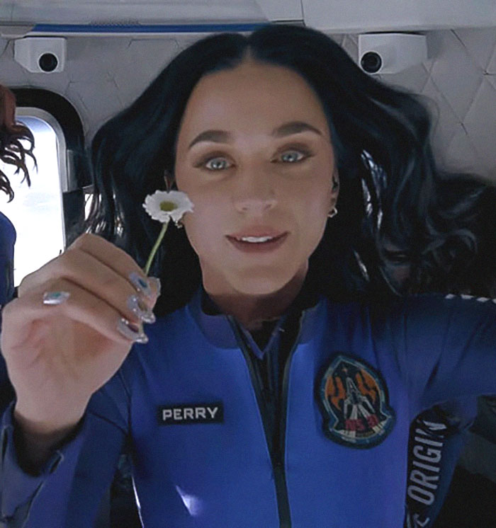 A woman in a space suit holds a flower, with "PERRY" on her uniform, highlighting tour debut and space theme. A woman in a space suit holds a flower, with "PERRY" on her uniform, highlighting tour debut and space theme.