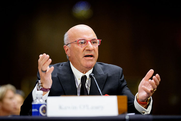 Kevin O’Leary speaking at a formal event, wearing a suit and red glasses, symbolizing his growing net worth and success.