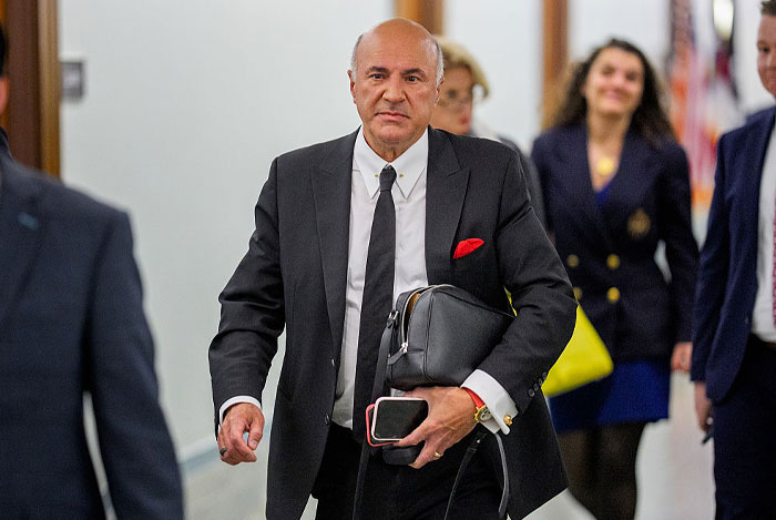 Kevin O’Leary in a black suit carrying a bag and phone, walking confidently, highlighting his net worth and business success.