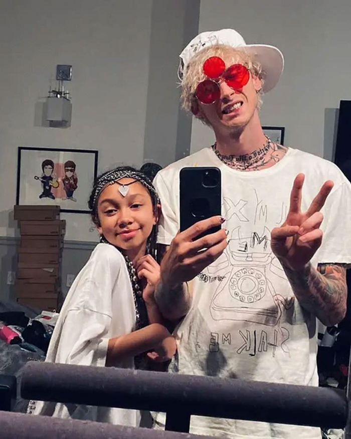 Machine Gun Kelly taking a selfie, wearing red glasses and a white hat, making a peace sign with a young girl. Machine Gun Kelly taking a selfie, wearing red glasses and a white hat, making a peace sign with a young girl.