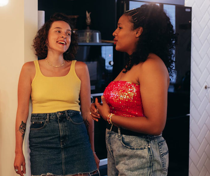 Two women in casual conversation in a kitchen setting, wearing summer outfits, smiling and enjoying each other's company. Two women in casual conversation in a kitchen setting, wearing summer outfits, smiling and enjoying each other's company.