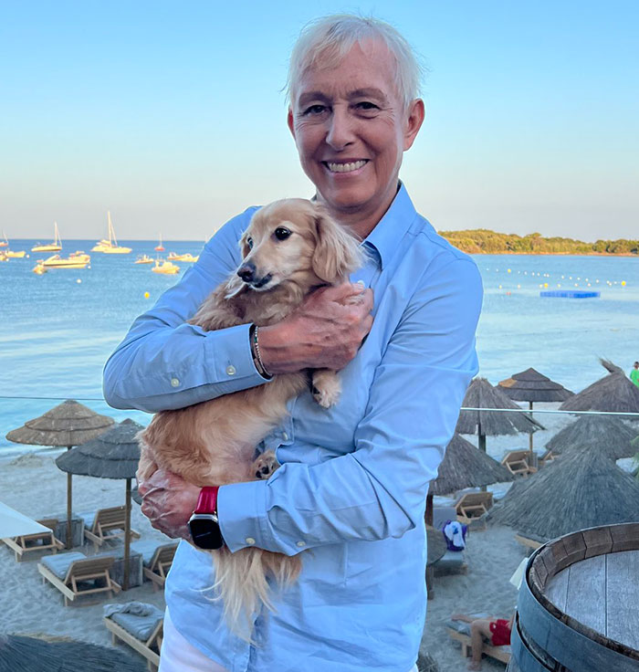 Smiling person in a blue shirt holding a dog by the beach, yachts in the background, emphasizing female athlete disqualified theme. Smiling person in a blue shirt holding a dog by the beach, yachts in the background, emphasizing female athlete disqualified theme.