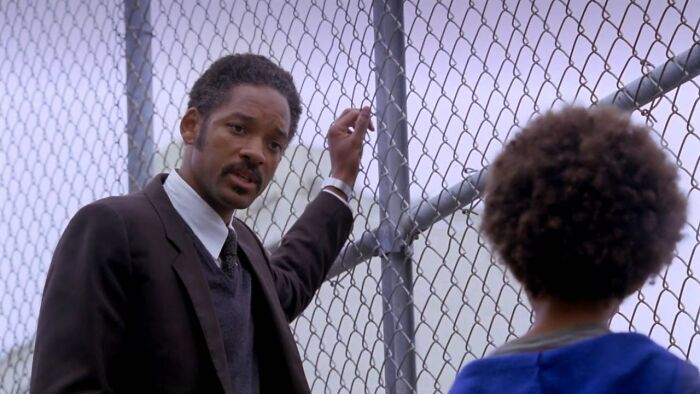 A man in a suit talking to a child by a chain-link fence, a touching movie scene that may cause tears.