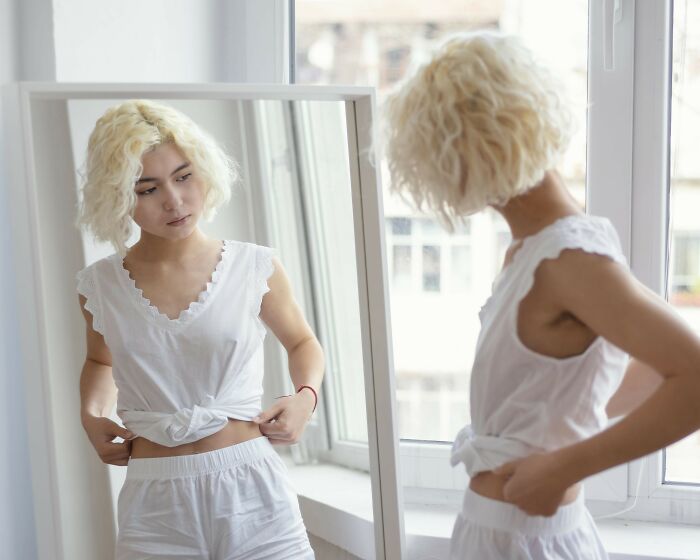 Woman in white outfit looking at her reflection, adjusting top in front of mirror, symbolizing dating reflections.