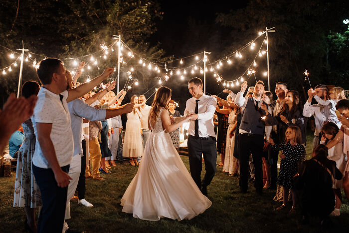 Couple dancing under string lights at an outdoor wedding reception surrounded by guests holding sparklers. Couple dancing under string lights at an outdoor wedding reception surrounded by guests holding sparklers.