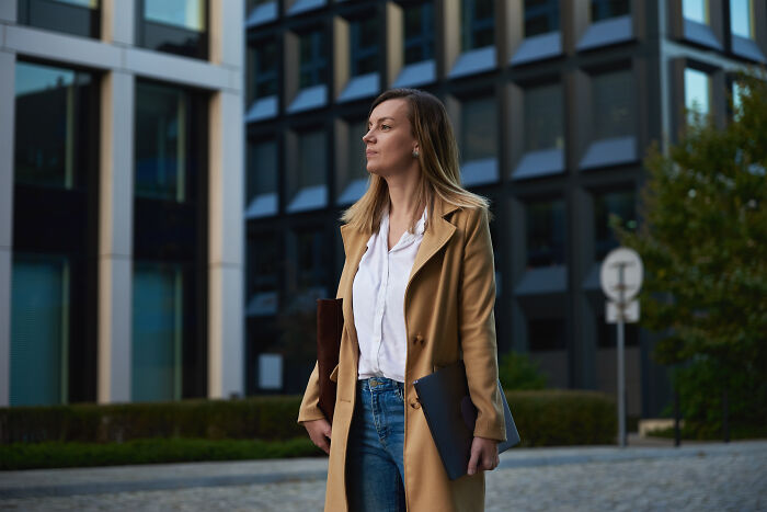 Woman holding a notebook, standing confidently in a city plaza, with modern buildings in the background.