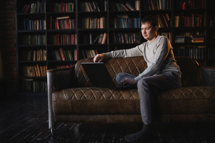 A person sitting on a brown sofa in a library, using a laptop, surrounded by colorful books.