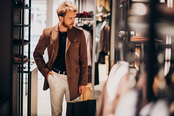 Man wearing a brown coat shopping in a clothing store, holding a bag amidst strangers.